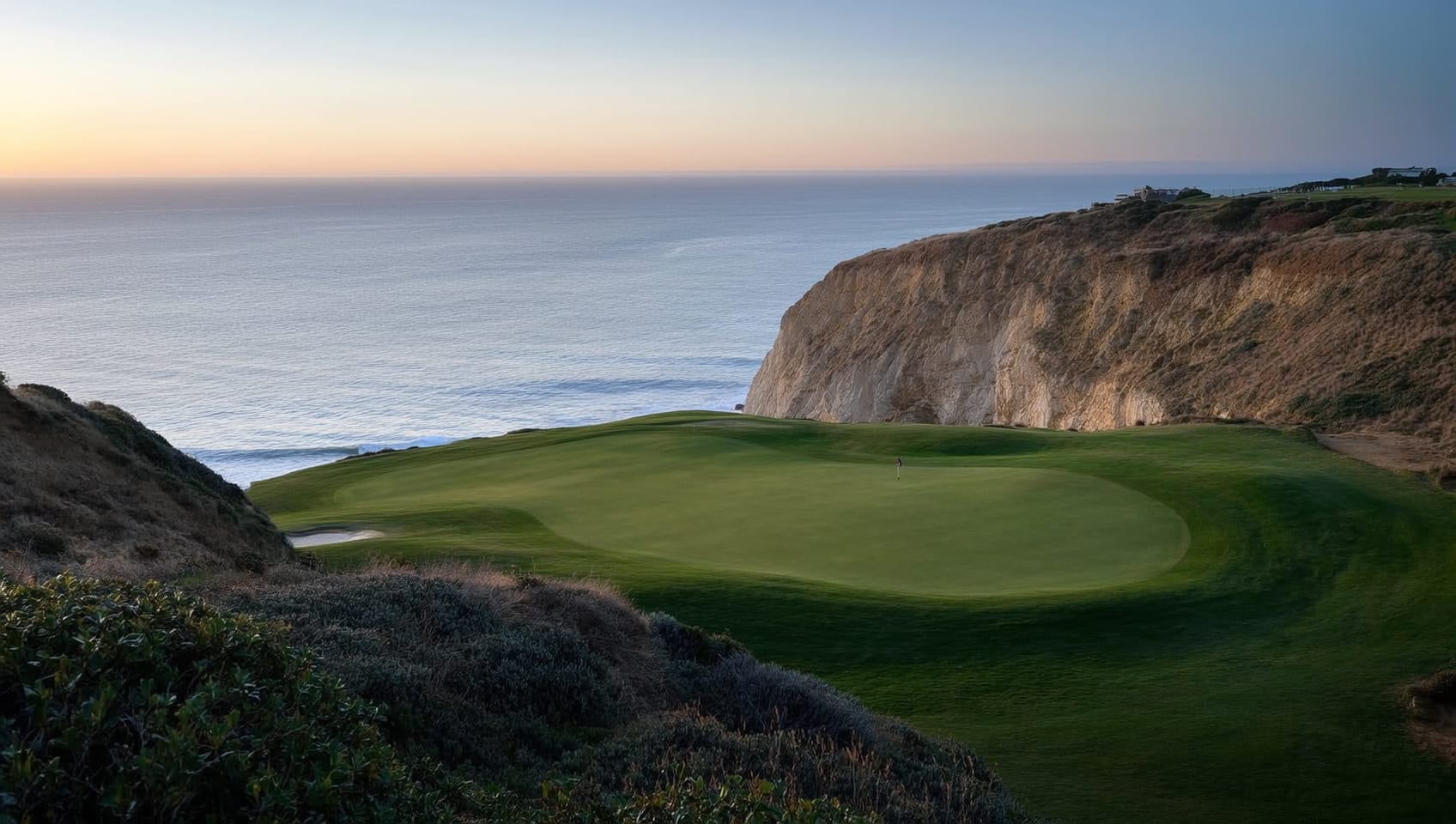 A cliffside golf green above the sea at dusk, framed by dramatic coastal terrain