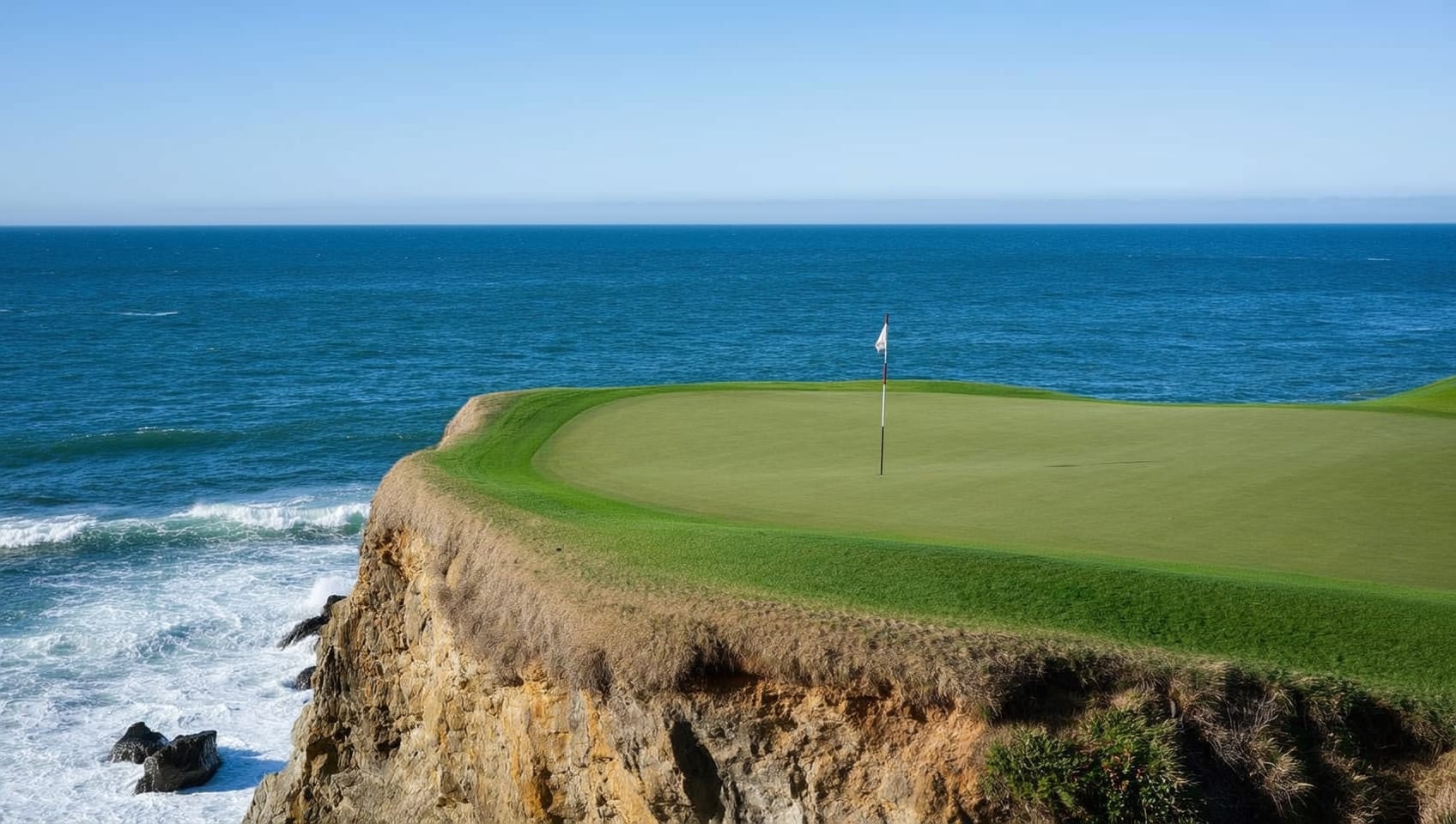 A golf green set on a grassy cliff above the Pacific Ocean, with a lone flagstick against blue water and sky