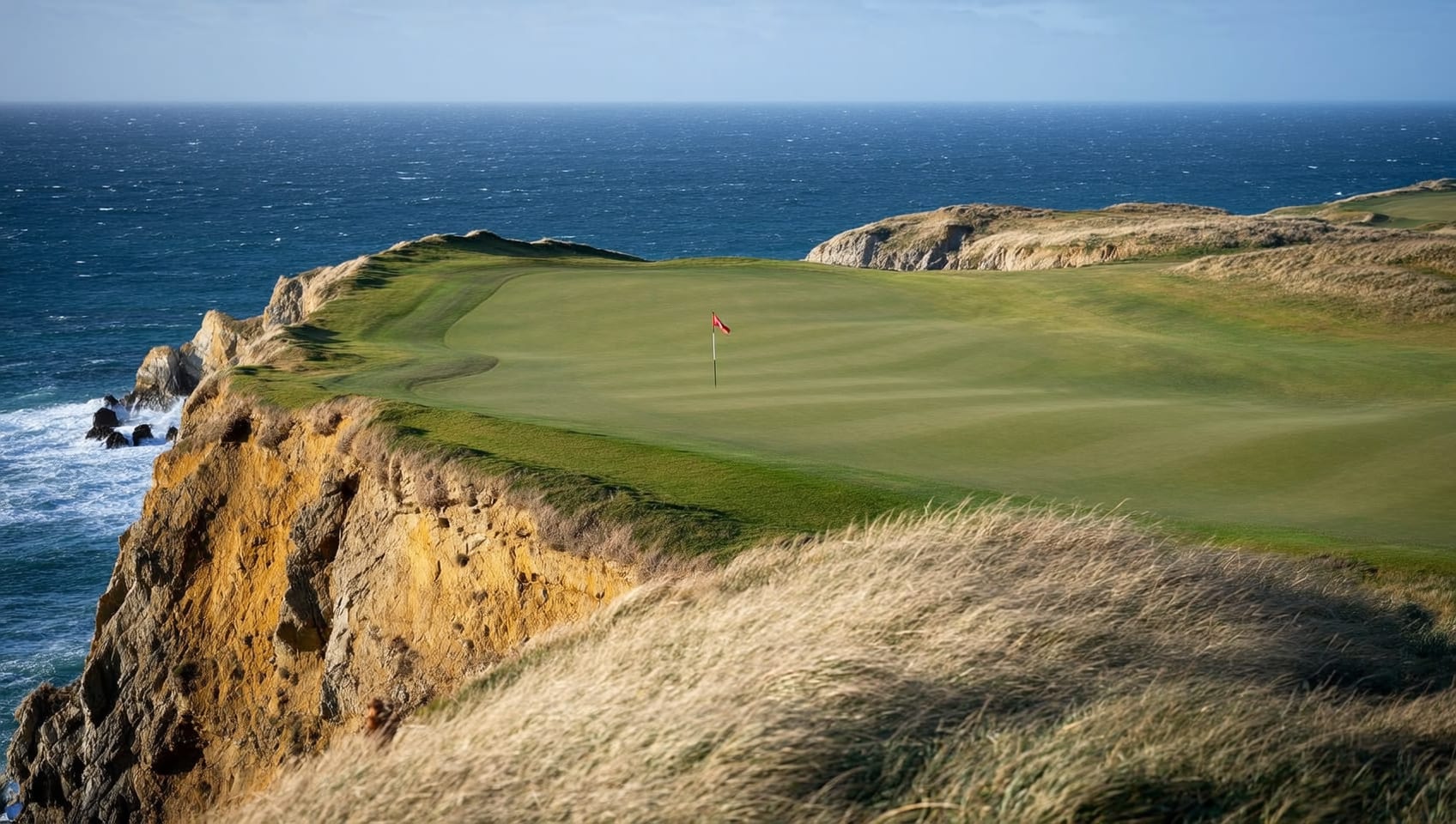 A cliff-top links fairway falling away toward the ocean, a lone red flag marking the green on a grassy headland