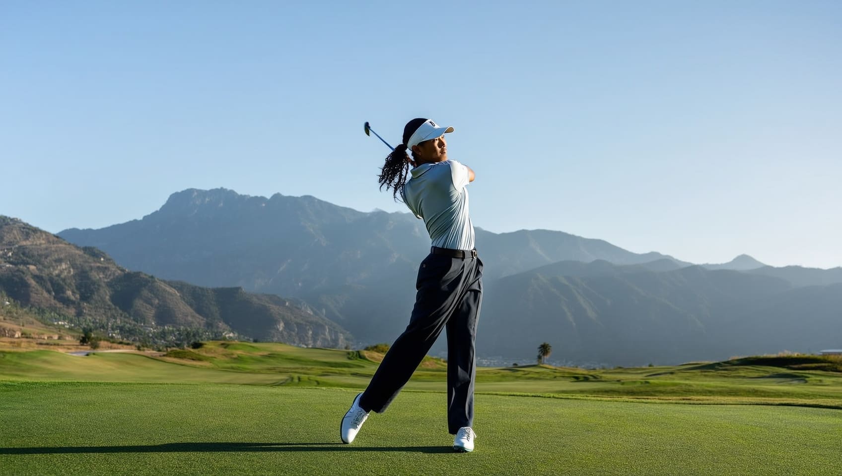 A golfer mid-swing on a mountain course with dramatic peaks rising behind a sunlit fairway